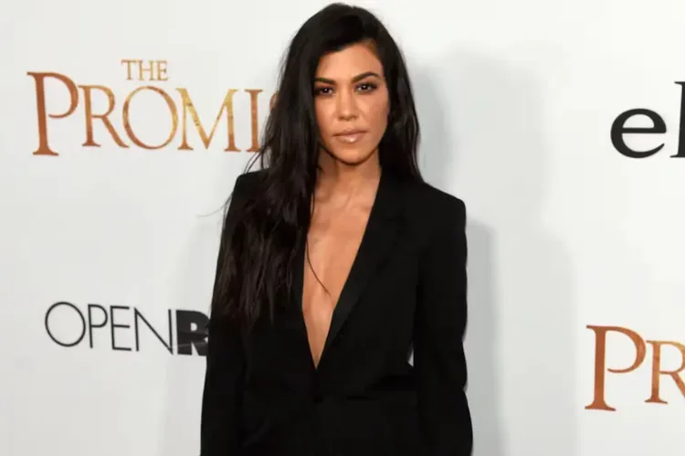 Kourtney Kardashian long dark hair a black coat wearing poses in front of in a white backdrop.