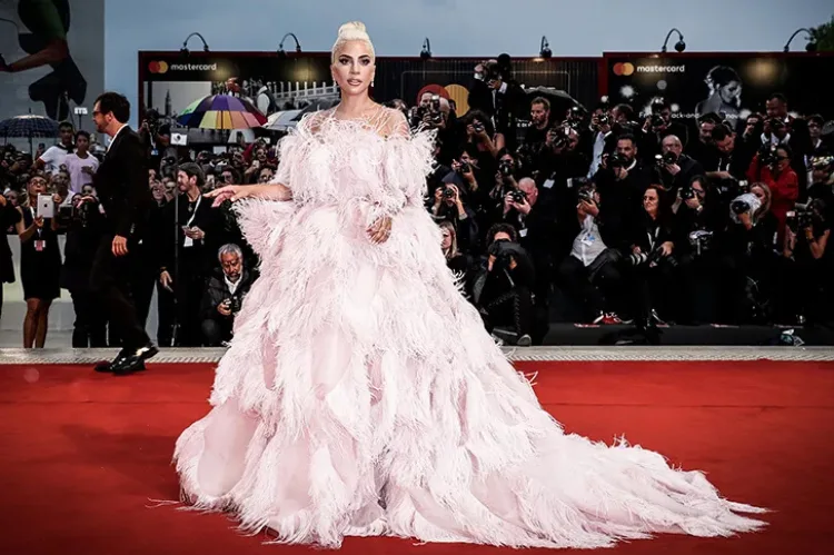 A celebritie in a dramatic pale pink feathered ball gown with a long train poses on the red carpet.